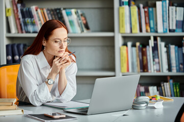 A redheaded female tutor is teaching an online lesson using a laptop at a desk.
