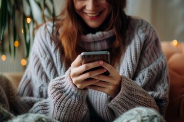 Fototapeta premium A happy young woman, sitting at home on the couch using her smartphone, smiling and looking at the screen while shopping online or working on social media apps. Social concepts, lifestyle