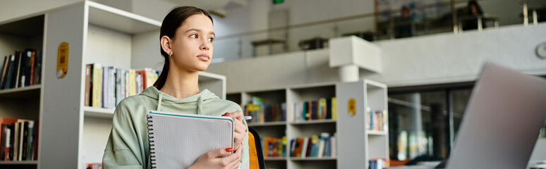 A teenage girl stands in front of a bookcase in a library, immersed in studying and doing homework on her laptop after school. © LIGHTFIELD STUDIOS