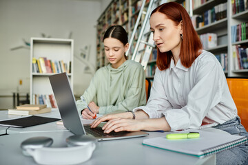Redhead tutor engages in after-school tutoring lesson with teenage girl in library setting, both focused on laptop.
