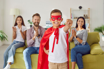 Portrait of a happy smiling child boy wearing red superhero costume standing in the living room at home with his family in background. Parents having fun with kids. Family time and leisure concept.