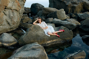 Serene woman in white dress sitting on rock by water, red shoes nearby