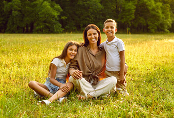 Happy woman with daughter and son sitting in the field on green grass and looking cheerful at camera. Smiling mother hugging her two children in the summer park. Family leisure concept.
