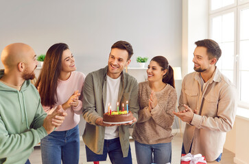 Smiling young happy man holding his cake with candles standing in the living room with cheerful joyful people friends congratulating him with a birthday at home party. Celebration concept.