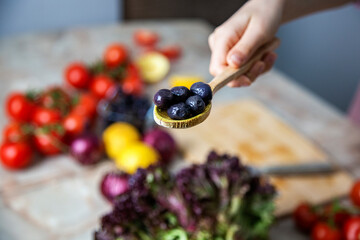 person holding a wooden spoon with black olives close-up against table with vegetables 