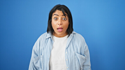 A surprised hispanic woman with wide eyes stands against an isolated blue background, embodying...