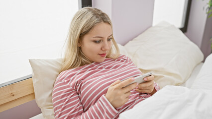 Blonde woman relaxing in bed with smartphone, evoking leisure, technology, and indoor lifestyle.