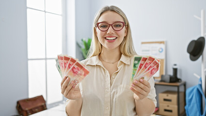 Blonde woman in glasses smiling while holding israeli shekels in a bright office interior.