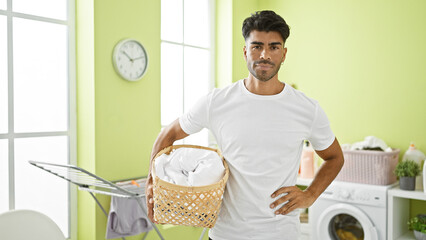 A handsome young man with a beard doing laundry in a bright green room, holding a basket and...