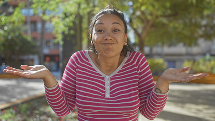 A young hispanic woman in a striped shirt gestures with a puzzled expression in a sunny park with...