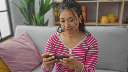 A young hispanic woman relaxes in her living room, focused on her smartphone, displaying everyday...