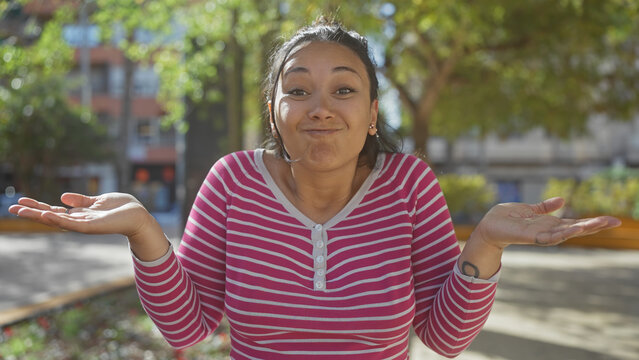 A young hispanic woman in a striped shirt gestures with a puzzled expression in a sunny park with blurred greenery in the background.
