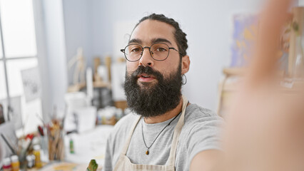 Hispanic man with beard in an art studio taking a selfie, portraying a creative and relaxed atmosphere.
