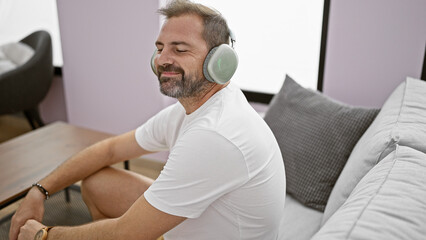 Obraz premium Relaxed middle-aged man with beard smiling, wearing headphones and white shirt, sitting on a couch indoors.