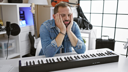 Hispanic man with grey hair in denim jacket looks exhausted at a keyboard in a modern music studio.