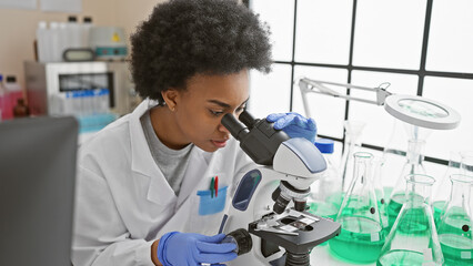 African american woman scientist using microscope in laboratory setting surrounded by equipment.