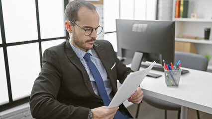 Fototapeta premium A focused businessman in glasses reads documents in a modern office setting, exemplifying professionalism and concentration.