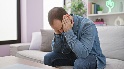 Fototapeta premium Bald stressed man sitting on a white sofa indoors holding head in hands with a blurred interior background.