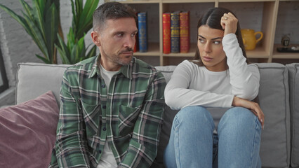 A hispanic couple appears troubled sitting together on a grey sofa indoors, showing tension and contemplation.