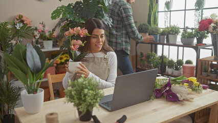 Woman florist in apron using laptop for video call in flower shop with man arranging plants in background