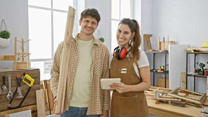 A smiling man and woman teamwork in a bright carpentry studio with woodworking tools.