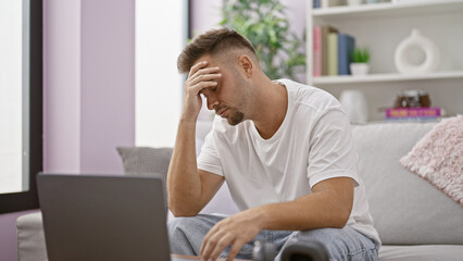A stressed young hispanic man with a beard sitting in a living room while using a laptop.