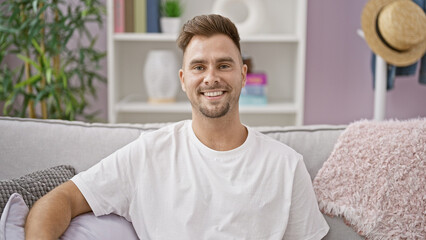 A handsome, young man with a beard sitting on a couch in a cozy living room, exuding casual relaxation and comfort.