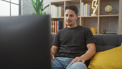 Handsome young hispanic man with a beard sitting on a sofa in a cozy living room at home, looking relaxed.