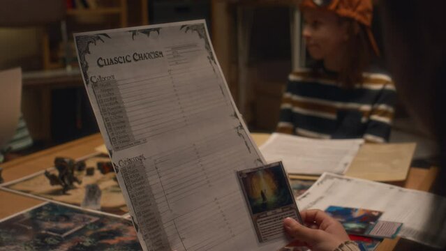 Over shoulder shot of anonymous kid holding character sheet and unique character card before starting playing board game with friends in evening indoors