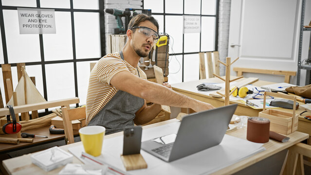 Handsome hispanic man with beard working on a laptop in a well-equipped carpentry workshop. - Powered by Adobe