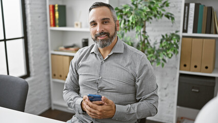 A mature hispanic man with a beard and grey hair smiles while holding a smartphone in a modern office setting.