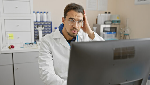 Worried young hispanic man in lab coat analyzing data on computer in hospital laboratory.