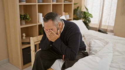 Obraz premium A mature hispanic man with a beard and grey hair sits distressed in a bedroom, covering his face with hands.