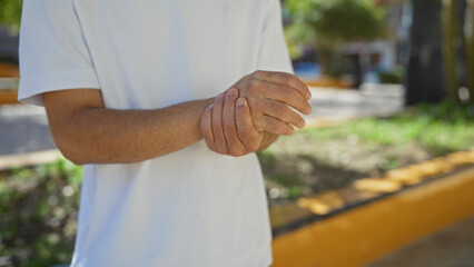 A young hispanic man in a white t-shirt holds his hands together in an outdoor urban park setting,...