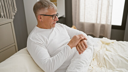Mature grey-haired man in white shirt relaxing on bed in a well-lit bedroom interior