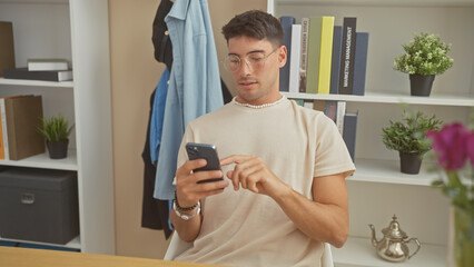 Handsome hispanic man in glasses using smartphone at home, seated by a table with a shelved background.