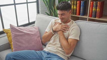 Handsome hispanic man smiling with hands on chest, seated on a gray sofa in a cozy living room.