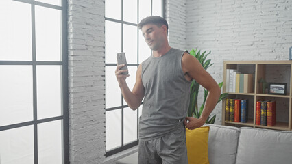 Man in casual grey activewear using smartphone in a sunny, modern apartment living room with sofa and bookshelf.