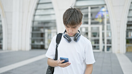 A young man with headphones around his neck checks his smartphone on a university campus.