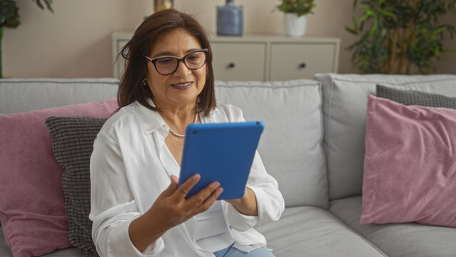 An elderly hispanic woman with short hair and glasses using a tablet, sitting on a couch in a living room at home, surrounded by cushions and indoor plants.
