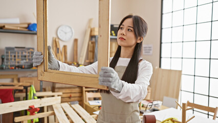 Chinese woman examines wooden frame thoughtfully in sunlit carpentry workshop.