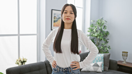 Confident asian woman standing in a modern living room with hands on hips and natural light flooding in