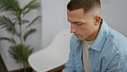 Fototapeta premium Handsome young man in a light blue shirt sitting thoughtfully in a modern indoor room with a blurred plant in the background
