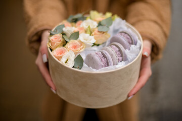 Girl in a brown jacket holds a cardboard box of charming flowers and delightful lilac macarons in her hands