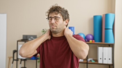 A young man experiencing neck pain in a clinic's rehabilitation room, portraying discomfort and the need for physical therapy.
