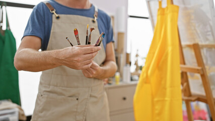 A man in an apron holds paintbrushes in a sunlit art studio, evoking creativity and artistic work.