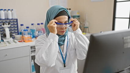 A woman in a hijab adjusts safety goggles in a laboratory setting, exemplifying professionalism and diversity.