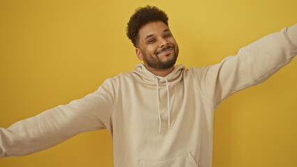 Smiling african american man in casual hoodie posing with arms wide open against a yellow background.