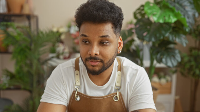 A contemplative african american man with a beard, wearing an apron, sits amidst lush greenery in a sunlit flower shop.