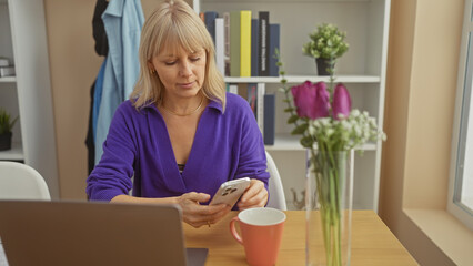 Fototapeta premium Blonde woman using smartphone at home office with laptop, flowers, and coffee mug